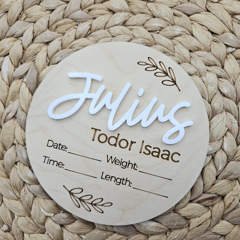 May include: A circular wooden birth announcement sign featuring the name Julius in raised white script lettering and the name Todor Isaac in engraved text. The sign includes engraved fields for Date, Time, Weight, and Length with leaf accents.