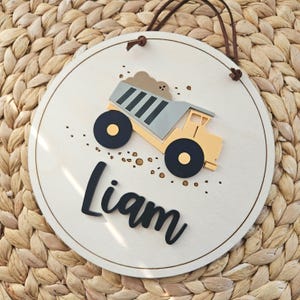 May include: A circular wooden sign with a light brown edge and a brown leather hanging cord. It features a yellow and grey dump truck with black wheels and the name "Liam" in black cursive. Small brown dots are scattered around the truck.