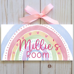 May include: A rectangular sign featuring a pastel watercolour rainbow design. The sign reads "Millie's Room" in pink script. A pink ribbon is tied at the top for hanging. The sign is set against a light wood background.