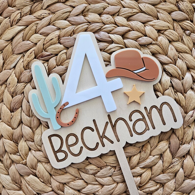 Peut inclure: D&eacute;coration de g&acirc;teau en bois sur le th&egrave;me du cow-boy. Elle pr&eacute;sente le chiffre 4 en blanc, un chapeau de cow-boy marron, un cactus bleu clair, un fer &agrave; cheval et le nom "Beckham" dans une police d&eacute;corative. Une petite &eacute;toile jaune est &eacute;galement incluse.