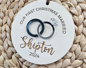 May include: A circular wooden Christmas ornament with the inscription "OUR FIRST CHRISTMAS MARRIED" and the name "Shipton" with the year "2024". It features two interlocking rings and a diamond. A brown leather cord is attached for hanging.