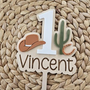 Peut inclure: Un cake topper en bois sur le th&egrave;me du cowboy. Le topper pr&eacute;sente un chapeau de cowboy marron, un cactus vert, un fer &agrave; cheval et le chiffre 1 en blanc. Le nom "Vincent" est &eacute;crit en police script.