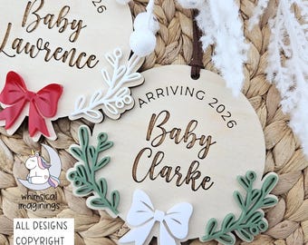 May include: Two circular wooden ornaments featuring the engraved text ARRIVING 2026 and Baby Lawrence or Baby Clarke. One ornament displays a red bow and white sprig, while the other features a white bow and two green sprigs, both with brown hanging cords.