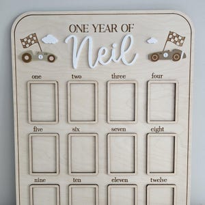 以下が含まれることがあります： 「ONE YEAR OF Neil」の文字が入った木製のマイルストーンボード。1から12までの数字が振られた12個の写真スロットがあります。レースカーと雲のモチーフで装飾されています。