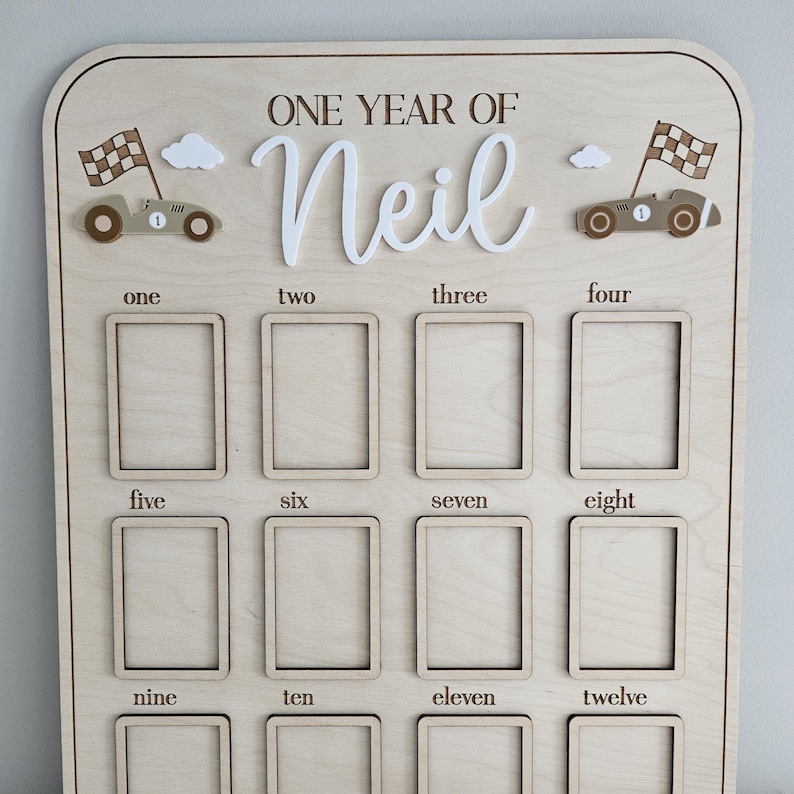 以下が含まれることがあります： 「ONE YEAR OF Neil」の文字が入った木製のマイルストーンボード。1から12までの数字が振られた12個の写真スロットがあります。レースカーと雲のモチーフで装飾されています。