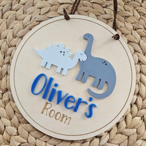 May include: Circular wooden sign featuring two dinosaur illustrations in light blue and grey. The sign displays the text "Oliver's Room" in blue and brown, with a brown leather hanging cord.