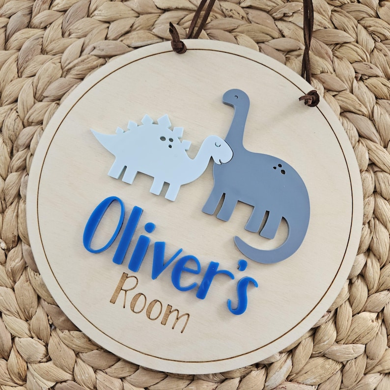 May include: Circular wooden sign featuring two dinosaur illustrations in light blue and grey. The sign displays the text "Oliver's Room" in blue and brown, with a brown leather hanging cord.