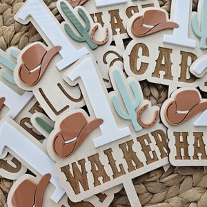 Peut inclure: Des d&eacute;corations de g&acirc;teau en bois sur le th&egrave;me du cow-boy. Les d&eacute;corations comprennent le chiffre "1" en blanc, des chapeaux de cow-boy marron, des cactus verts et le nom "WALKER" dans une police western. Les d&eacute;corations sont &eacute;parpill&eacute;es sur une surface tiss&eacute;e.