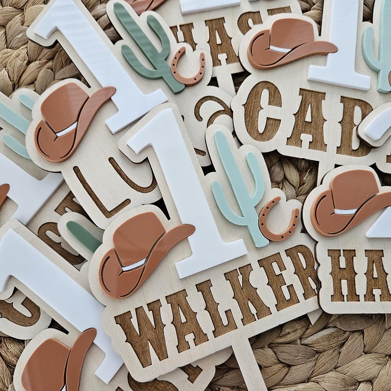 Peut inclure: Des d&eacute;corations de g&acirc;teau en bois sur le th&egrave;me du cow-boy. Les d&eacute;corations comprennent le chiffre "1" en blanc, des chapeaux de cow-boy marron, des cactus verts et le nom "WALKER" dans une police western. Les d&eacute;corations sont &eacute;parpill&eacute;es sur une surface tiss&eacute;e.