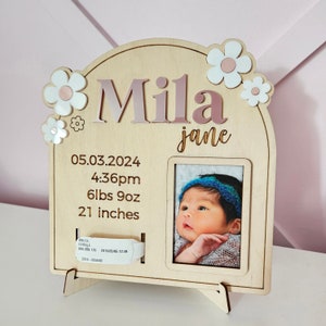 May include: Wooden baby photo frame with the name "Mila" in pink letters, adorned with flower accents. Includes baby's birth details: date, time, weight (6lbs 9oz), and length (21 inches). A photo of the baby is displayed.