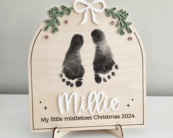 May include: Wooden Christmas ornament with baby footprints, the name "Millie", and the text "My little mistletoes Christmas 2024". The ornament has a bow and greenery design at the top.