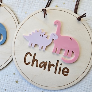 May include: Round wooden sign with a light wood finish, featuring a pink and a light pink dinosaur design. The name "Charlie" is engraved in a brown font. The sign has a brown leather cord for hanging.