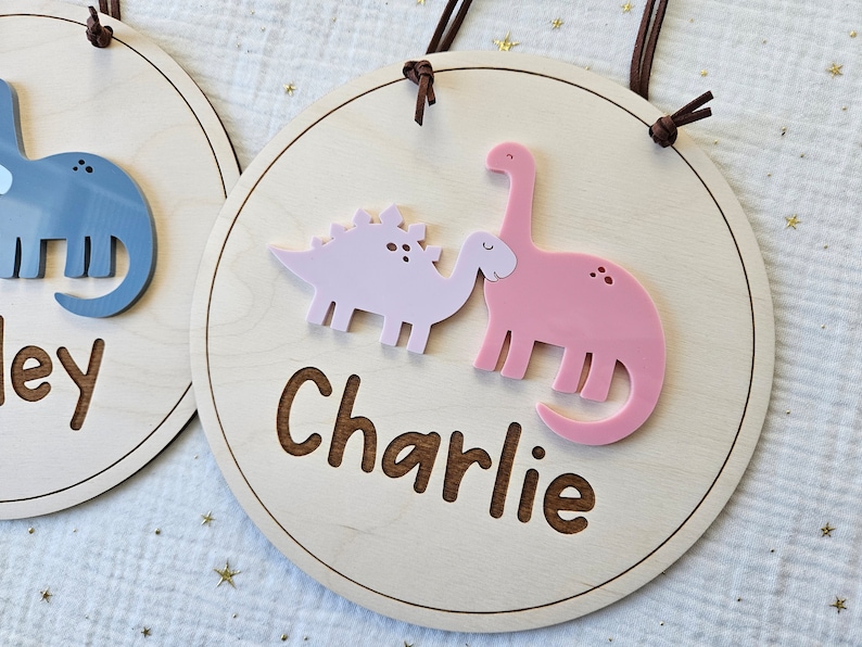 May include: Round wooden sign with a light wood finish, featuring a pink and a light pink dinosaur design. The name "Charlie" is engraved in a brown font. The sign has a brown leather cord for hanging.