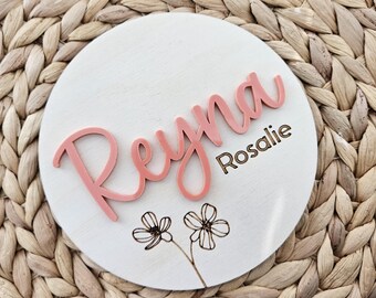 Peut inclure: Panneau rond en bois blanc avec le nom "Reyna" en lettres roses en relief. Le nom "Rosalie" est gravé en dessous. Deux fleurs esquissées sont en bas. Le panneau est posé sur un fond tissé.
