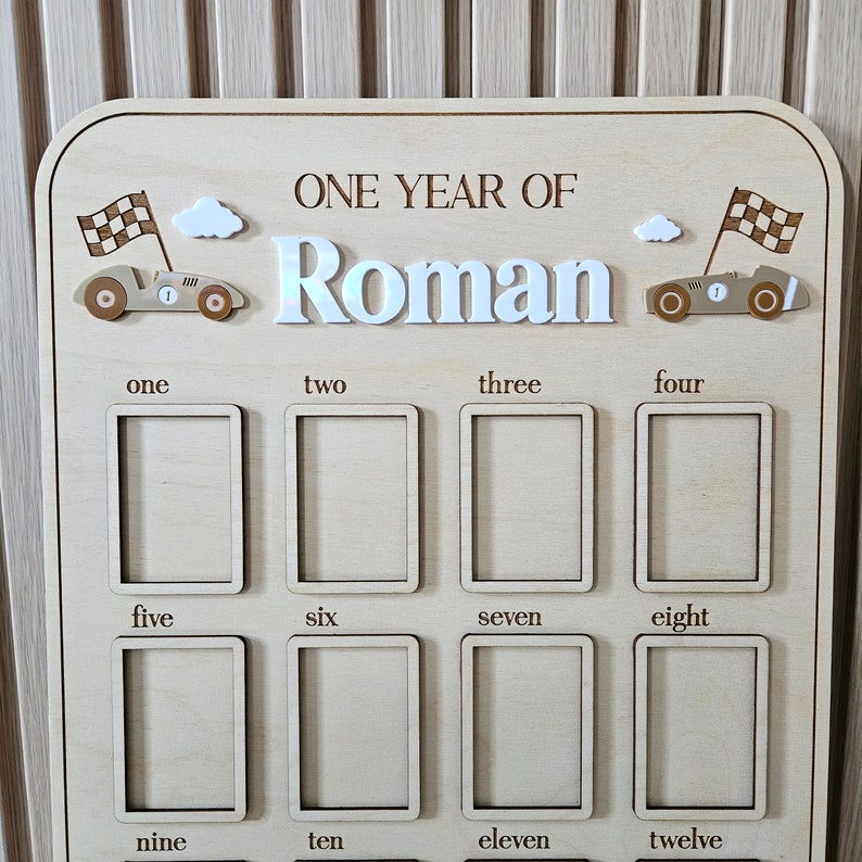以下が含まれることがあります： 「ONE YEAR OF Roman」の文字が入った木製のマイルストーンボード。レースカーとチェッカーフラッグのデザインで、1から12までの写真スペースがあります。「Roman」という名前は、白く虹色のフォントで表示されています。