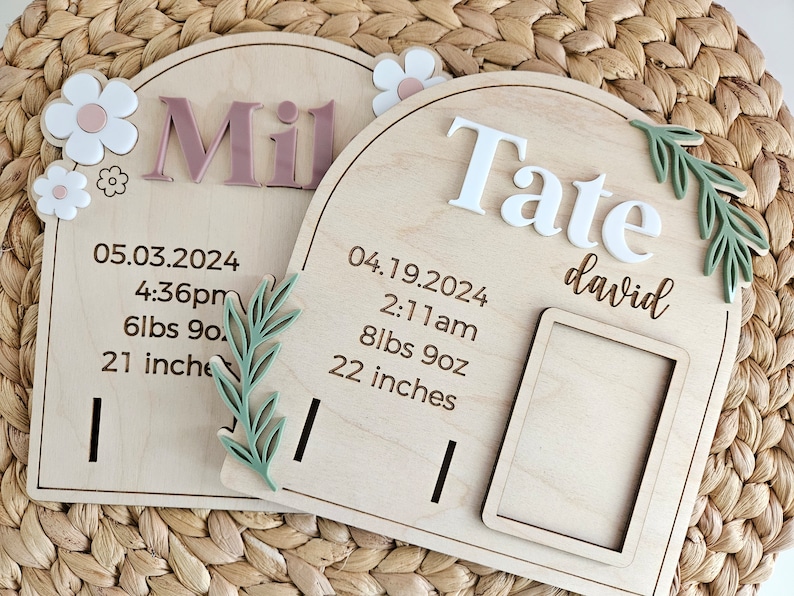 May include: Two wooden birth announcement plaques with arched tops. One has the name "Mil" in pink letters, the other "Tate" in white. Both include birth details: date, time, weight (lbs oz), and length (inches). Floral and leaf accents.