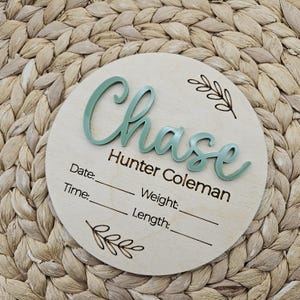 May include: A circular wooden birth announcement sign featuring the name Chase Hunter Coleman in raised sage green script lettering. The board includes laser-engraved text for Date, Time, Weight, and Length, accompanied by two small leaf illustrations.