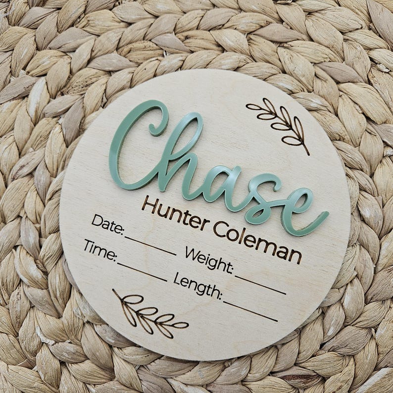 May include: A circular wooden birth announcement sign featuring the name Chase Hunter Coleman in raised sage green script lettering. The board includes laser-engraved text for Date, Time, Weight, and Length, accompanied by two small leaf illustrations.