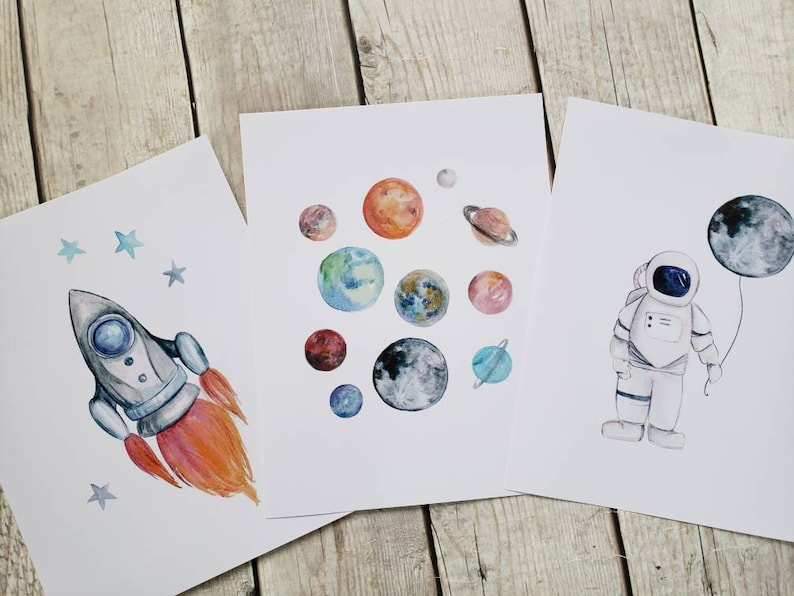 May include: Three watercolor art prints featuring space themes. One print shows a rocket ship with orange flames and blue stars. Another displays colorful planets. The third depicts an astronaut holding a moon-shaped balloon.