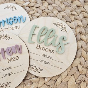 May include: Three circular wooden plaques for birth announcements. Each plaque features a laser-engraved leaf design, a name in raised acrylic lettering, and printed lines for weight and length. The names are Ellis, Harrison, and a partial name.