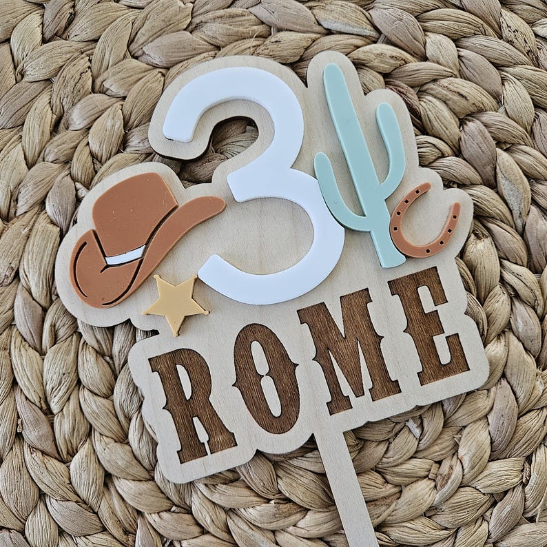 Peut inclure: Un cake topper en bois sur le th&egrave;me du cowboy. Le design comprend le chiffre 3 en blanc, un chapeau de cowboy marron, une &eacute;toile, un cactus et le nom "ROME" sur une pancarte en bois. Le topper est con&ccedil;u pour une f&ecirc;te d'anniversaire.