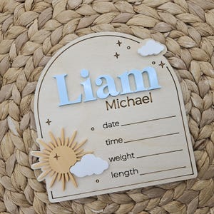 May include: An arched wooden birth announcement sign featuring the name Liam Michael in raised light blue lettering. The plaque includes a 3D sun and white clouds, with engraved lines for date, time, weight, and length, plus star accents.