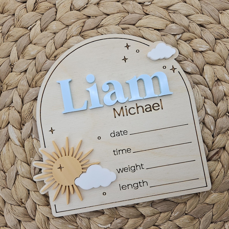 May include: An arched wooden birth announcement sign featuring the name Liam Michael in raised light blue lettering. The plaque includes a 3D sun and white clouds, with engraved lines for date, time, weight, and length, plus star accents.