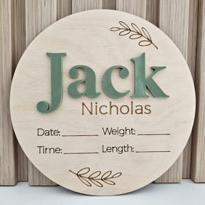 May include: A circular wooden birth announcement sign featuring the name Jack Nicholas in raised sage green acrylic lettering. The board includes laser-engraved text for Date, Weight, Time, and Length with blank lines, accented by two engraved leaf sprigs at the top and bottom.