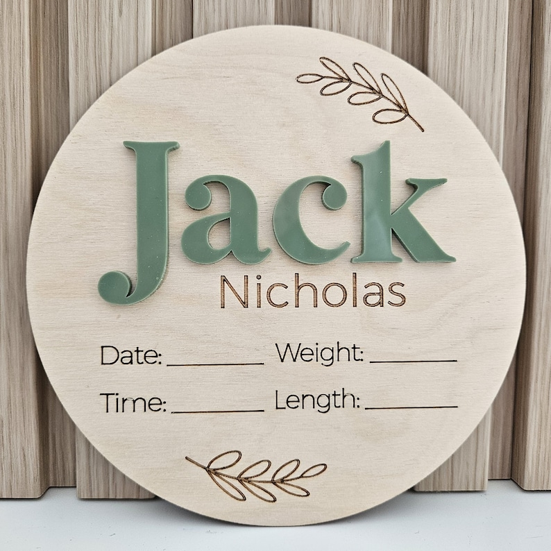 May include: A circular wooden birth announcement sign featuring the name Jack Nicholas in raised sage green acrylic lettering. The board includes laser-engraved text for Date, Weight, Time, and Length with blank lines, accented by two engraved leaf sprigs at the top and bottom.