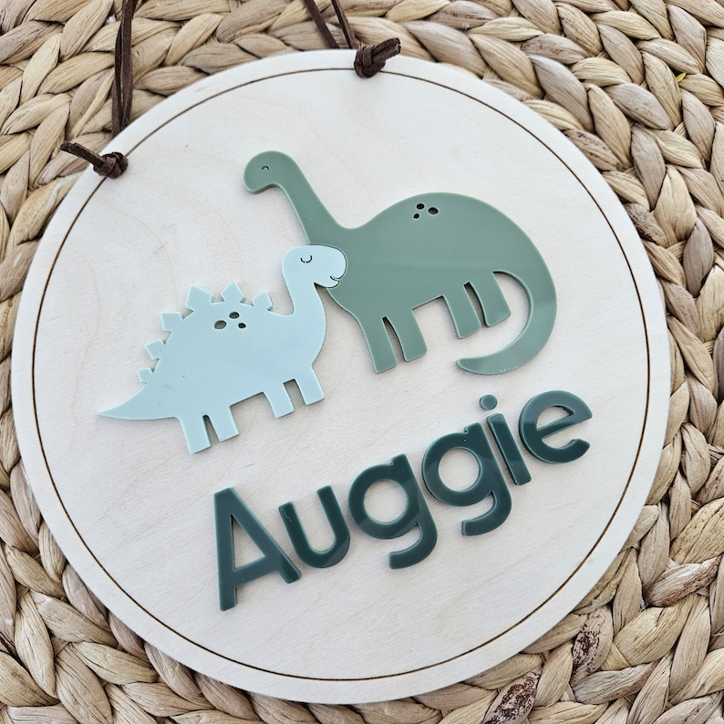 May include: Circular wooden sign with a light wood grain. It features two dinosaur cutouts in light blue and green, with the name "Auggie" in green lettering. Includes a brown leather hanging cord.