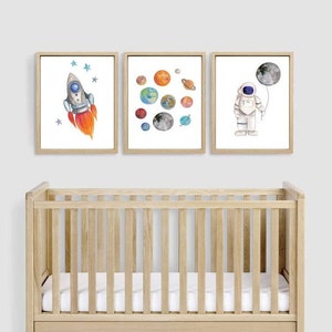 May include: Three framed watercolor prints with a space theme. One shows a rocket ship with orange flames, another depicts planets, and the third features an astronaut holding a moon balloon. The prints are displayed above a wooden crib.