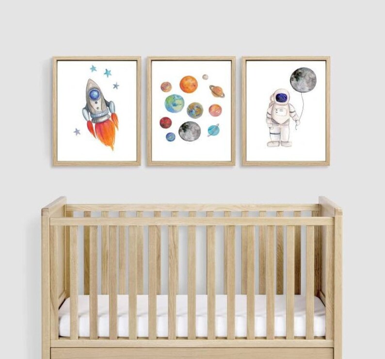 May include: Three framed watercolor prints with a space theme. One shows a rocket ship with orange flames, another depicts planets, and the third features an astronaut holding a moon balloon. The prints are displayed above a wooden crib.