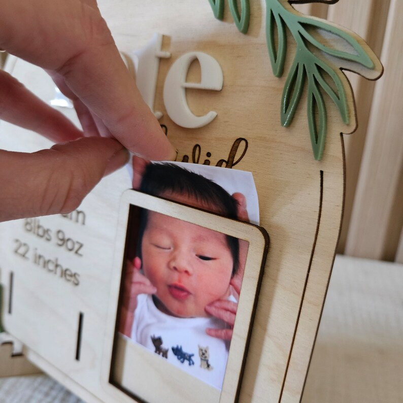 May include: A wooden baby photo frame with a picture of a newborn baby. The frame has the word "date" in white letters and a green leaf design. The baby's birth details are also included on the frame.