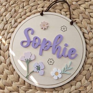 May include: A circular wooden sign featuring the name "Sophie" in lavender script, adorned with floral embellishments in lavender, pink, and white. A brown leather cord is attached for hanging.