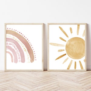 May include: Two framed watercolor prints. One features a rainbow in shades of pink and tan. The other depicts a sun with golden yellow rays. Both are on a white background, with light brown wooden frames. The art has a minimalist, bohemian style.