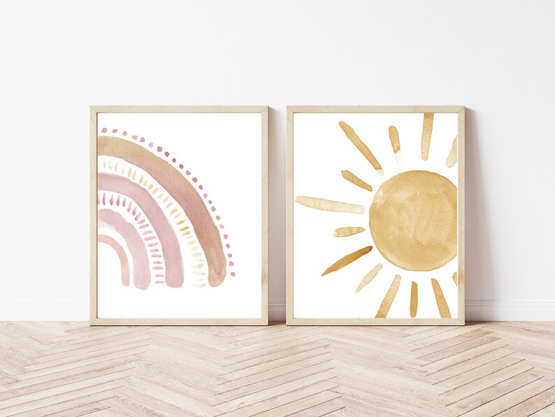 May include: Two framed watercolor prints. One features a rainbow in shades of pink and tan. The other depicts a sun with golden yellow rays. Both are on a white background, with light brown wooden frames. The art has a minimalist, bohemian style.