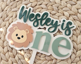 May include: A cake topper featuring a layered design with a tan lion face and the dark green text "Wesley is" above the light green text "one". The topper is constructed from multiple layers of material and includes a white stick base.