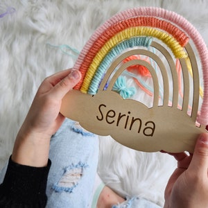 May include: A wooden rainbow-shaped wall hanging with the name "Serina" engraved on a cloud-shaped base. The rainbow features pastel colors of pink, orange, yellow, and blue yarn. A decorative item for a nursery or child's room.