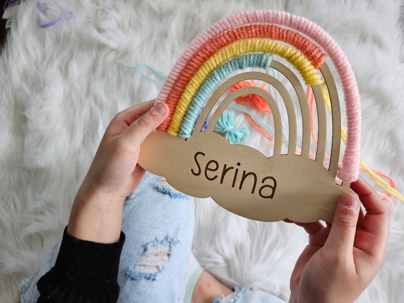May include: A wooden rainbow-shaped wall hanging with the name "Serina" engraved on a cloud-shaped base. The rainbow features pastel colors of pink, orange, yellow, and blue yarn. A decorative item for a nursery or child's room.