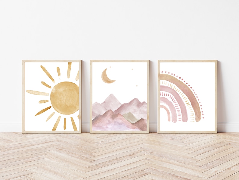 May include: Set of three framed watercolor art prints. The prints feature a sun, mountains with a moon, and a rainbow, all in soft, warm tones of gold, pink, and brown. The frames are a light wood color.