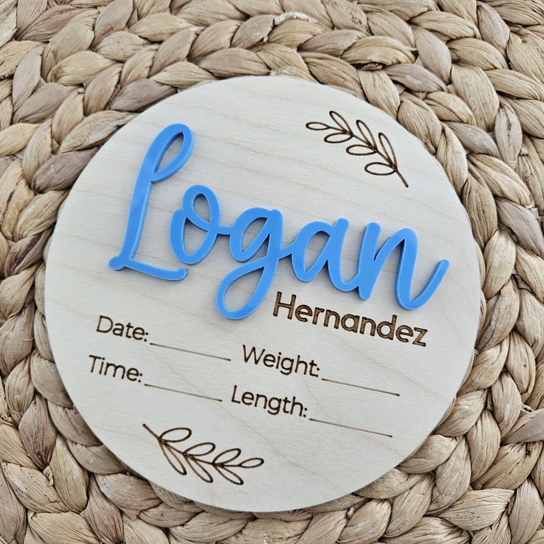 May include: A circular wooden plaque featuring the name Logan Hernandez in raised blue script lettering. The plaque includes laser-engraved text for Date, Weight, Time, and Length, accompanied by decorative leaf illustrations at the top and bottom.