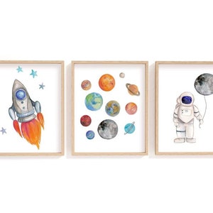 May include: Three framed watercolor prints with a space theme. One features a rocket ship with orange flames and blue stars. Another shows planets. The third depicts an astronaut holding a moon balloon.