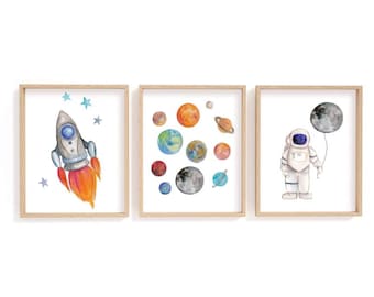 May include: Three framed watercolor prints with a space theme. One features a rocket ship with orange flames and blue stars. Another shows planets. The third depicts an astronaut holding a moon balloon.