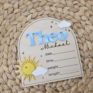 May include: An arched wooden birth announcement sign featuring the name Theo in raised blue letters and Michael in script. It includes a yellow sun, white clouds, and engraved lines for date, time, weight, and length, accented with small stars.