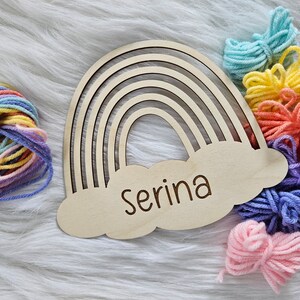 May include: Wooden rainbow-shaped wall decor with the name "Serina" engraved on a cloud. Colorful yarn bundles in shades of blue, yellow, orange, pink, and purple are arranged around the rainbow, creating a craft or nursery theme.