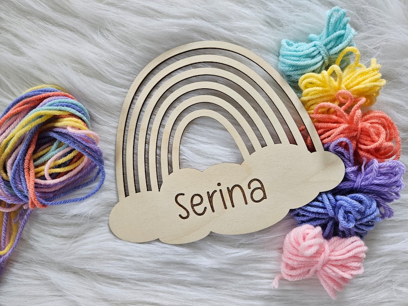 May include: Wooden rainbow-shaped wall decor with the name "Serina" engraved on a cloud. Colorful yarn bundles in shades of blue, yellow, orange, pink, and purple are arranged around the rainbow, creating a craft or nursery theme.