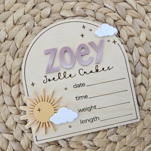 May include: An arched wooden birth announcement sign featuring the name ZOEY and Joelle Crakes in raised lettering. The plaque includes a yellow sun, white clouds, and star icons, with printed lines for date, time, weight, and length details.