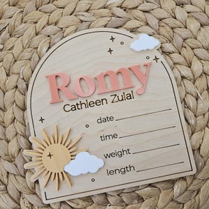 May include: An arched wooden birth announcement sign featuring the name Romy Cathleen Zulal in raised coral lettering. The plaque includes white acrylic clouds, a yellow sun, and engraved lines for date, time, weight, and length details.