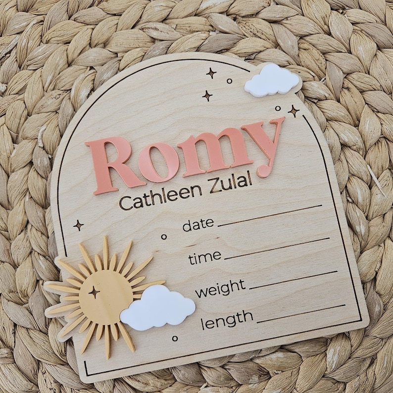 May include: An arched wooden birth announcement sign featuring the name Romy Cathleen Zulal in raised coral lettering. The plaque includes white acrylic clouds, a yellow sun, and engraved lines for date, time, weight, and length details.