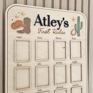 May include: A light-colored wooden board featuring the text Atley's First Rodeo, decorated with a cowboy hat, boot, cactus, horseshoe, and star icons. It includes twelve rectangular frames labeled one through twelve for photo displays.